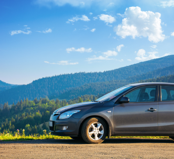 A grey car parked on a dirt road with green forested mountains and a blue sky with clouds in the background.