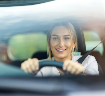 A young woman with blonde hair smiles while driving a car, seen through the front windshield. She is holding the steering wheel with both hands and wearing a seatbelt.