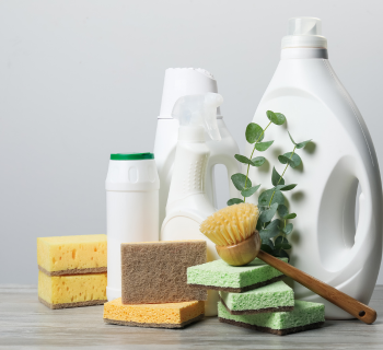 Various cleaning supplies, including plastic bottles, sponges, a scrub brush, and a sprig of eucalyptus, are arranged on a light wooden surface with a neutral background.