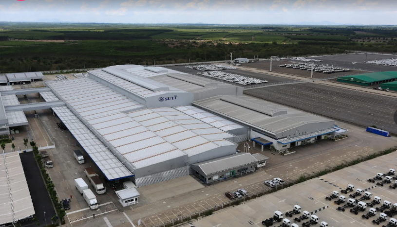 An aerial view of a large industrial facility with white rooftops, marked as "Siam Toyota," surrounded by trucks, cars, and vast parking lots, with green fields and mountains in the background.