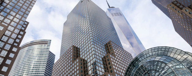 Several modern skyscrapers with glass facades rise into a partly cloudy sky, including the tall One World Trade Center in the background. The buildings reflect light and display a mix of geometric shapes and patterns.