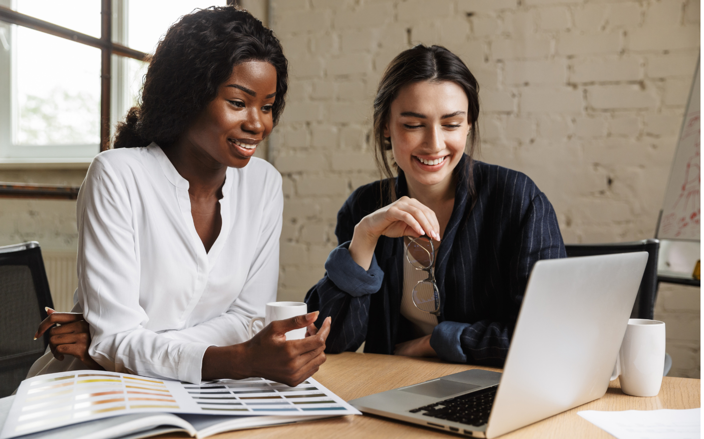 Two women sit at a desk, smiling and looking at a laptop screen featuring Therefore Business Central. One holds a coffee mug and color swatch booklet, while the other holds her glasses, suggesting a collaborative work environment.