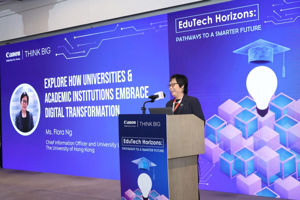A woman stands at a podium speaking at an event. Behind her, a large screen displays the title "Explore How Universities & Academic Institutions Embrace Digital Transformation" and event branding for "EduTech Horizons.